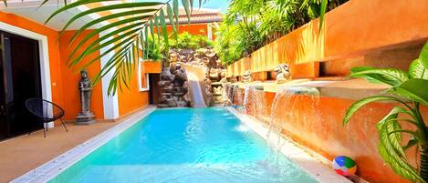 A swimming pool with a waterfall feature, surrounded by orange walls and tropical plants.
