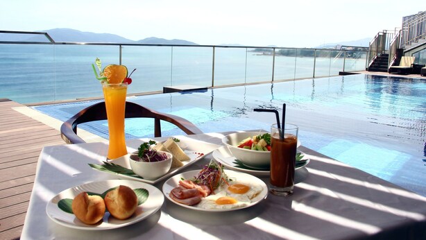 A dining table with a meal and drinks by a poolside with a view of the sea and mountains. - King Town Grand Hotel & Wedding Center (Bac Nha Trang)