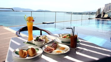 Una mesa de comedor con comida y bebidas junto a una piscina con vista al mar y a las montañas.