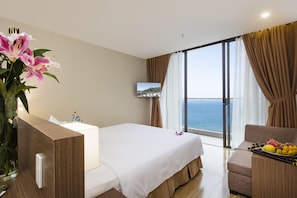 A hotel room with a bed, a desk, a chair, a TV, and a view of the sea.