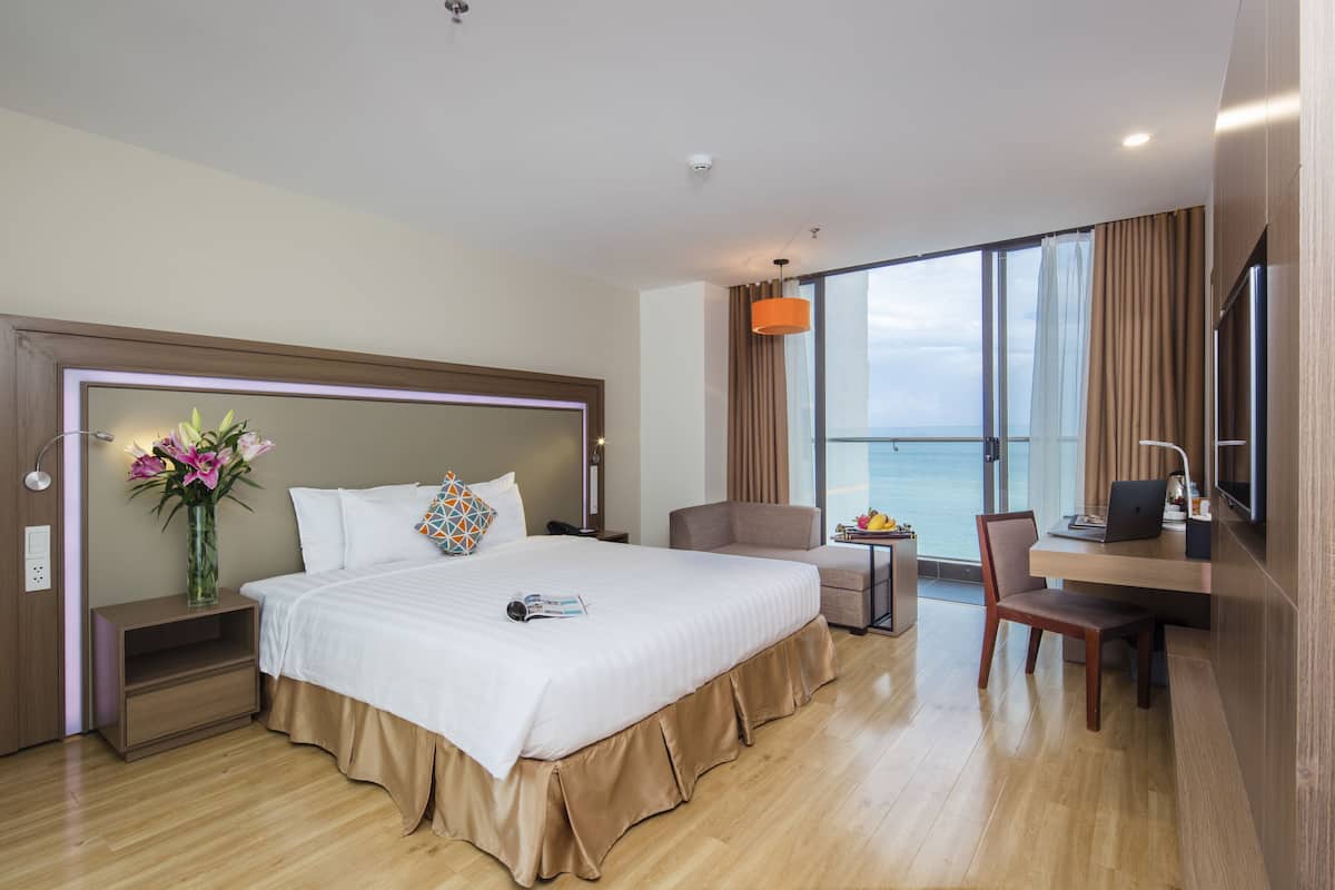 A modern hotel room with a large bed, a desk, and a view of the sea.