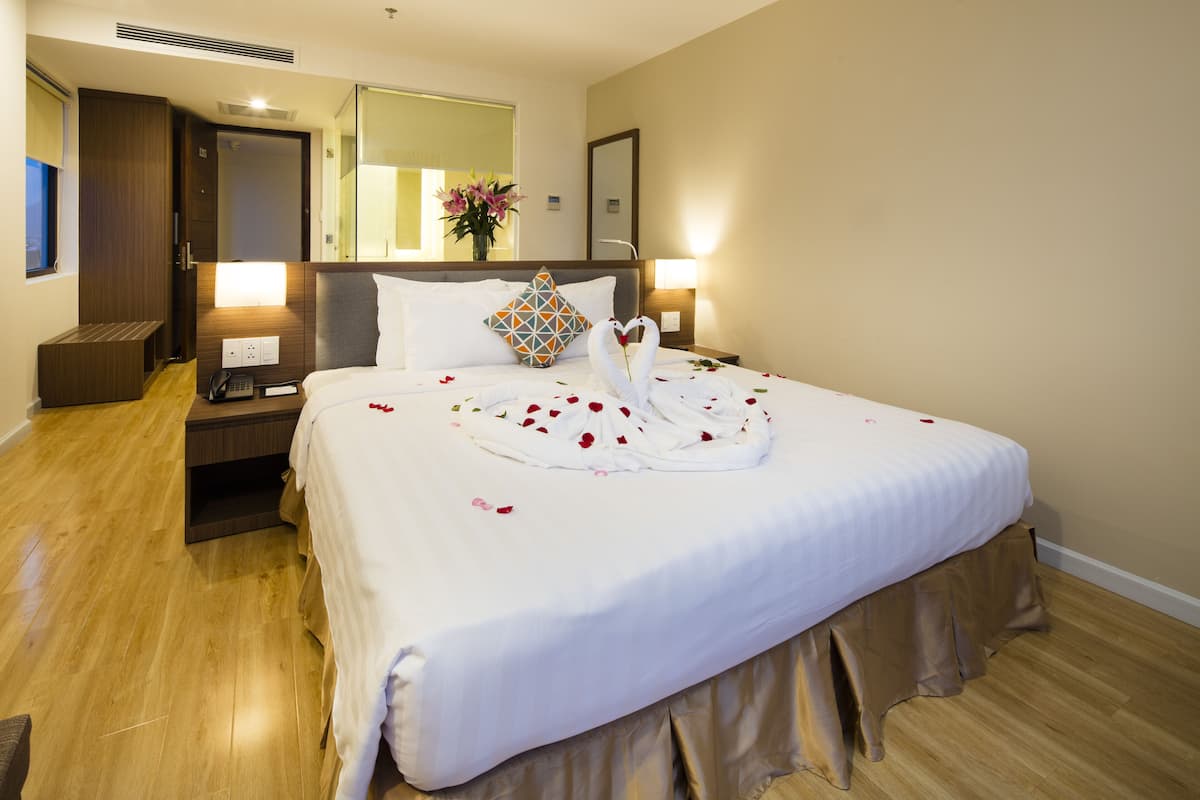A hotel room with a neatly made bed, a bedside table with a lamp, a nightstand, a wooden bench, and a large mirror on the wall.