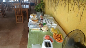 A buffet table with various food items, including bread, pastries, and sliced fruits, set up in a room with wooden chairs and a yellow wall. - Beach Villa Lanka (Tangalle)