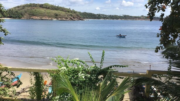A beach view with a boat on the water, a fenced area with plants, and a distant hill. - Beach Villa Lanka (Tangalle)