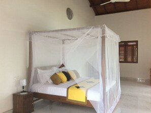 A four-poster bed with a mosquito net, a bedside table with a lamp, and a window with wooden shutters.