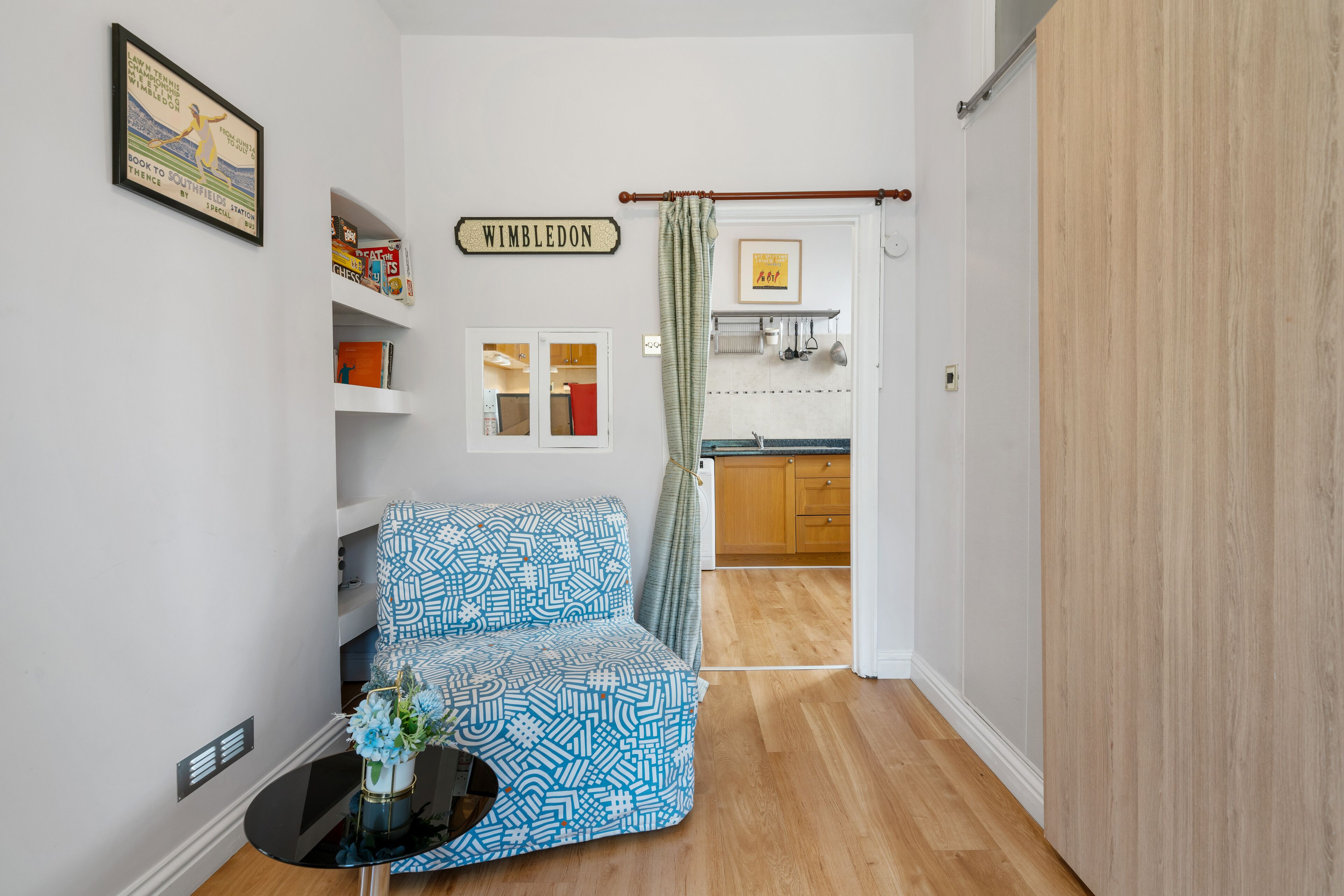 A compact living space with a patterned sofa, a small table with a vase, and a kitchen visible through an open door.
