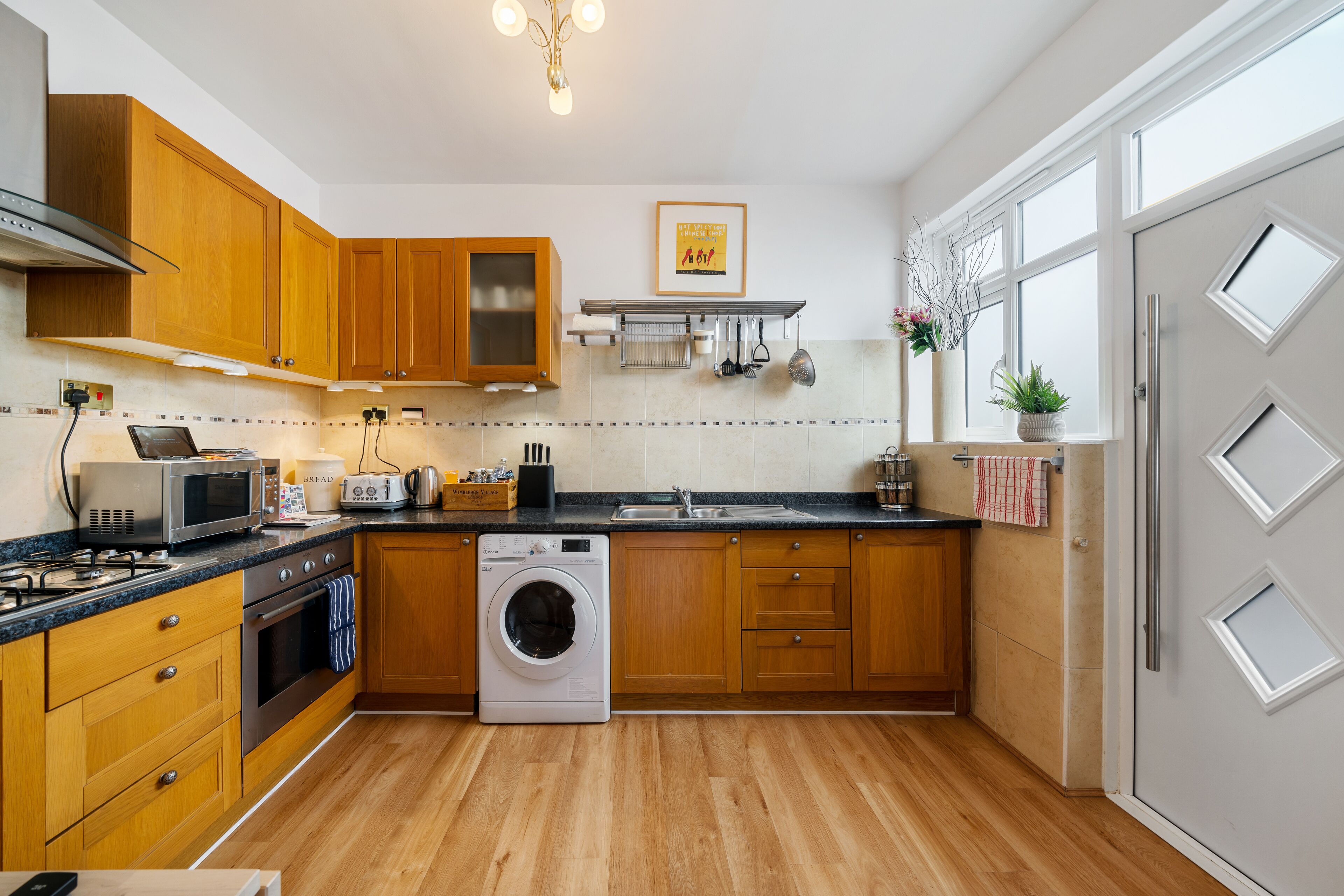 A modern kitchen with wooden cabinets, a built-in oven, a microwave, a washing machine, and a large window providing natural light.