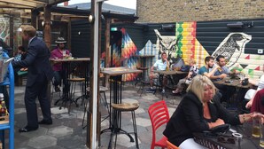 Un café en plein air avec des tables et des chaises en bois, un mur de briques orné de peintures murales colorées, et une personne qui sert des boissons.