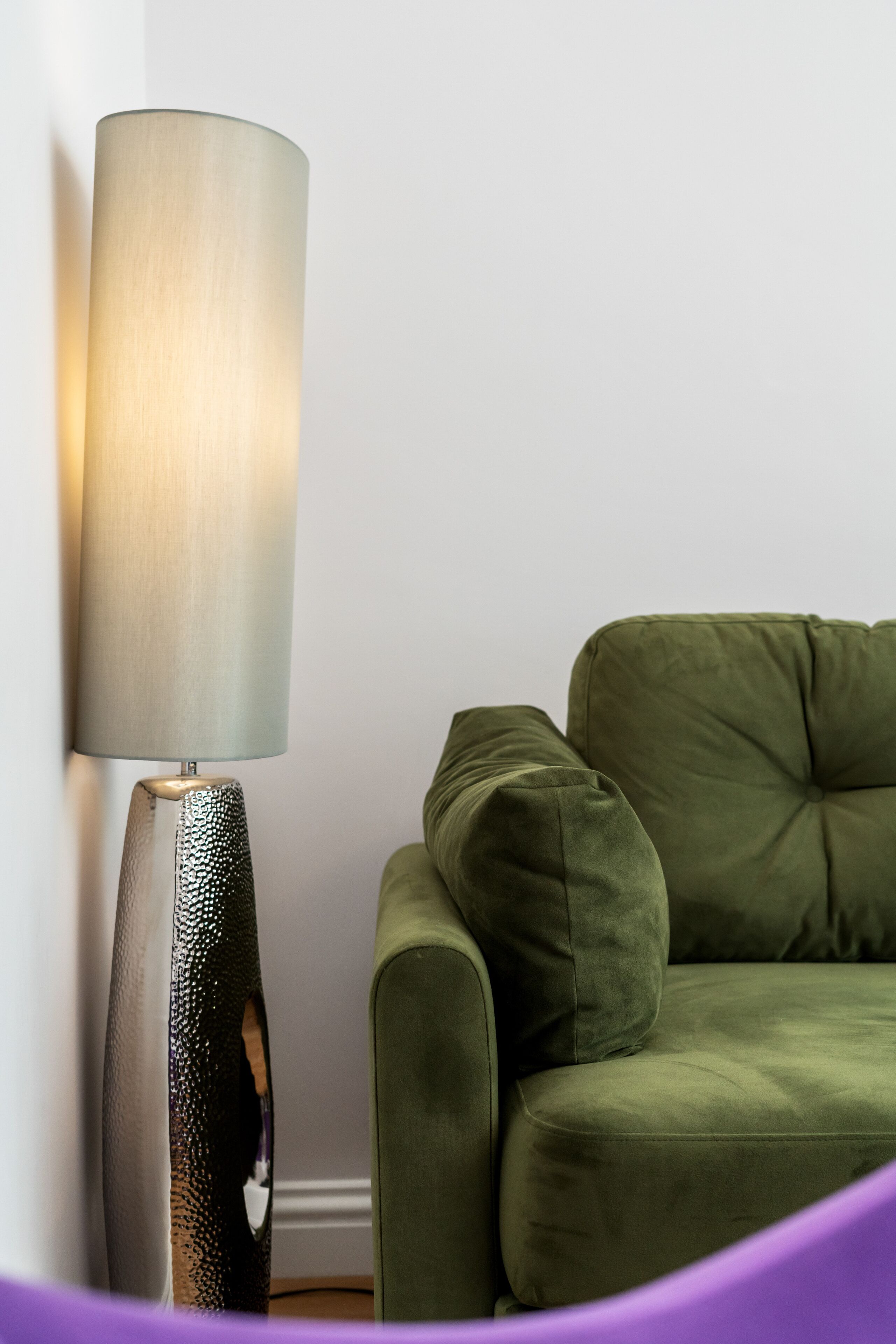 Eine moderne Lampe mit strukturierter Basis und zylindrischem Schirm, neben einem dunkelgrünen, gepolsterten Sofa vor einer weißen Wand.
