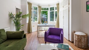 Une pièce avec un canapé vert, un fauteuil violet, une table à manger avec des chaises, une grande fenêtre avec des rideaux et un panier en osier.