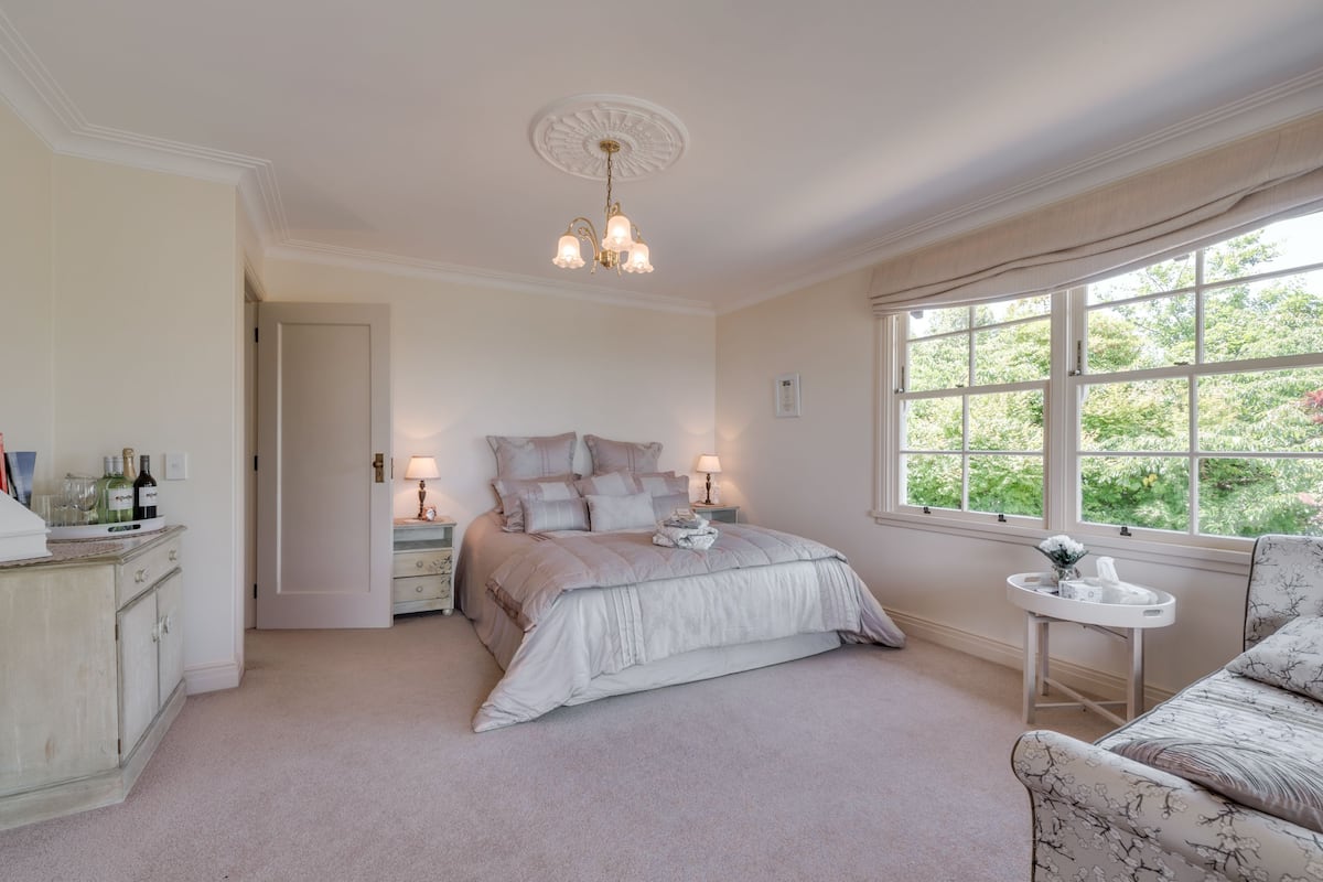 A bedroom with a large bed, bedside tables, a chandelier, a window with a view of greenery, and a floral-patterned chair.