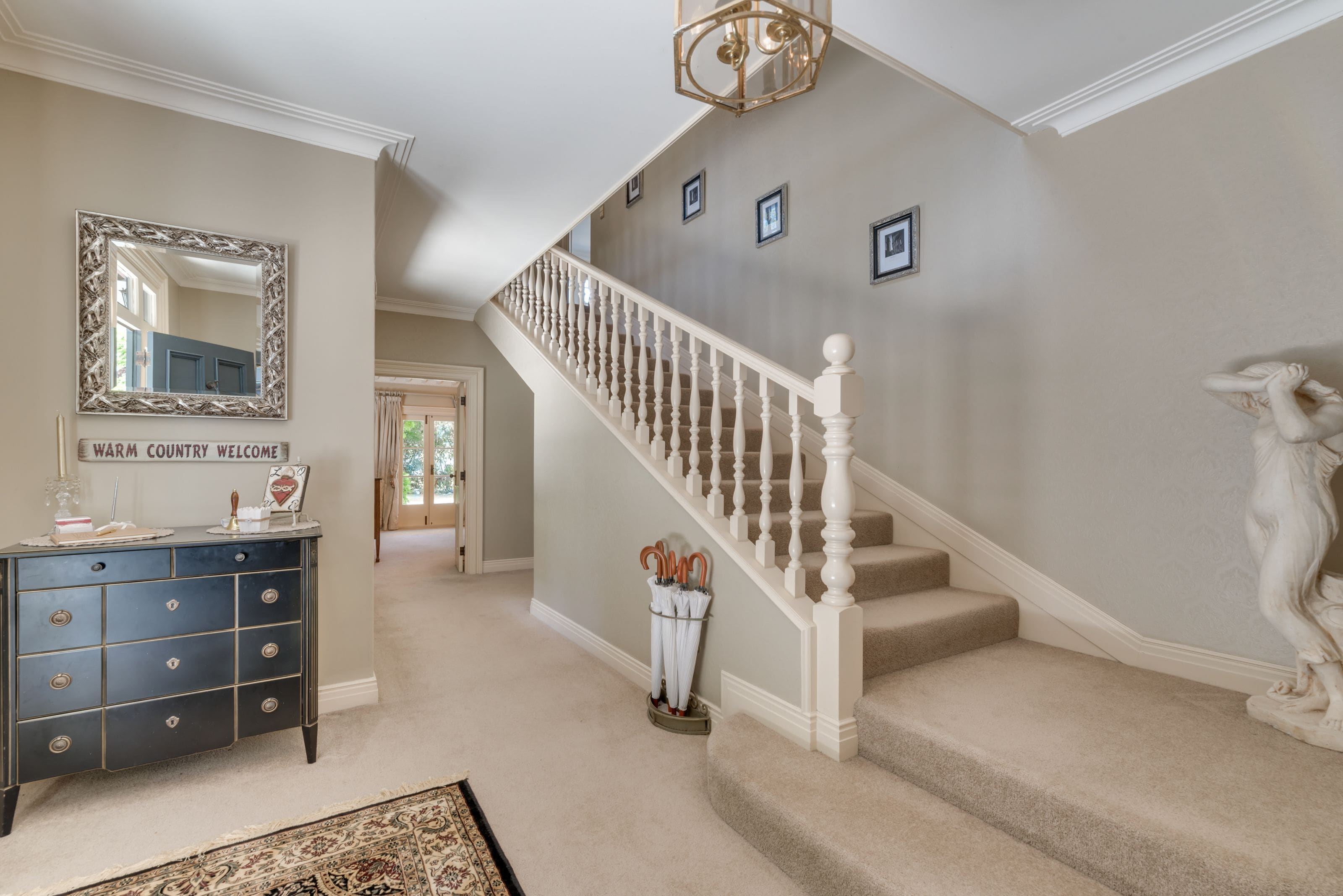 A grand staircase with a white balustrade leading to a landing. A dark wooden dresser with a mirror and a sign reading "WARM COUNTRY WELCOME" is visible.
