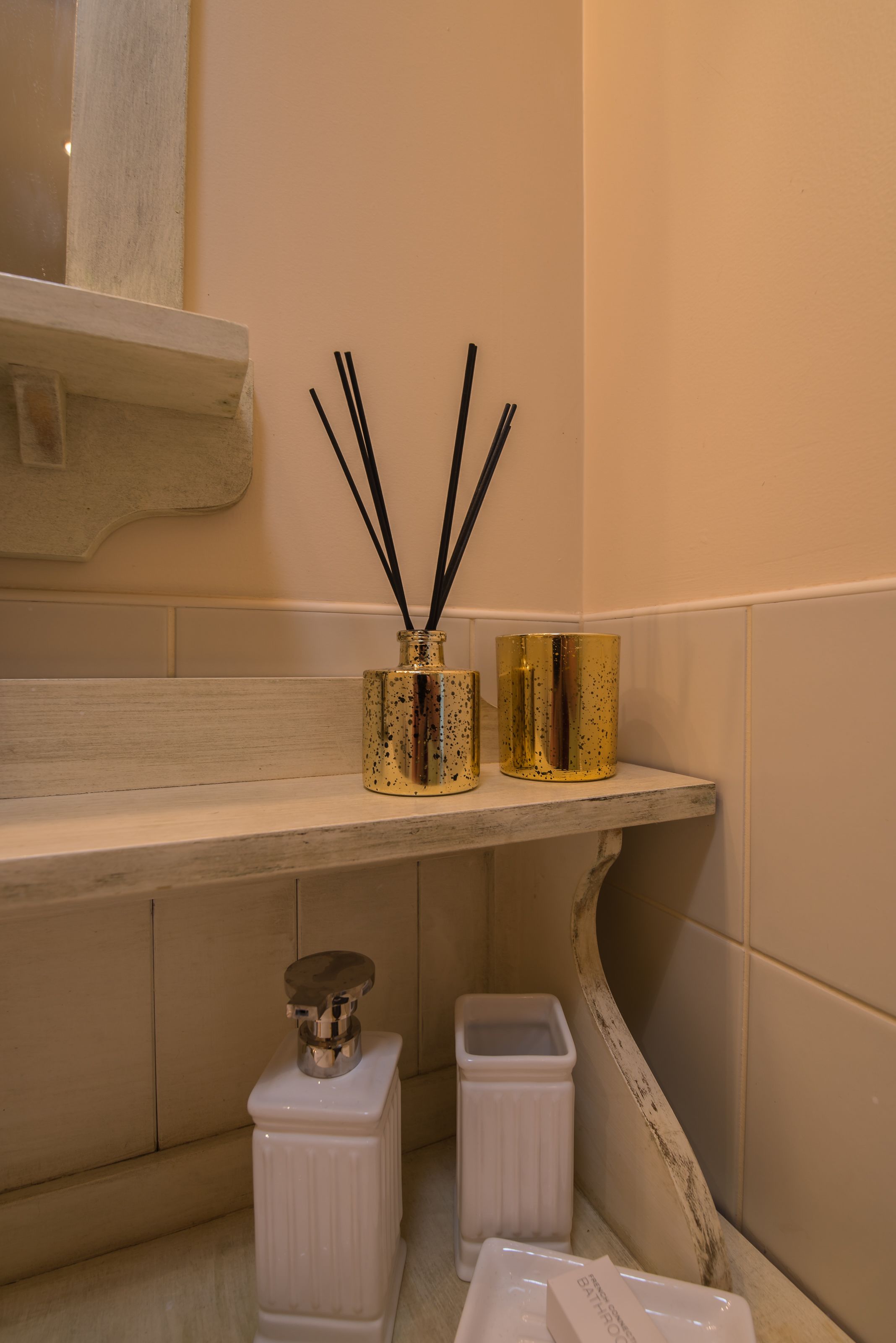 Un baño con pared de azulejos color beige, un lavabo blanco, un espejo y una estantería con dos recipientes dorados y palitos de incienso negros.