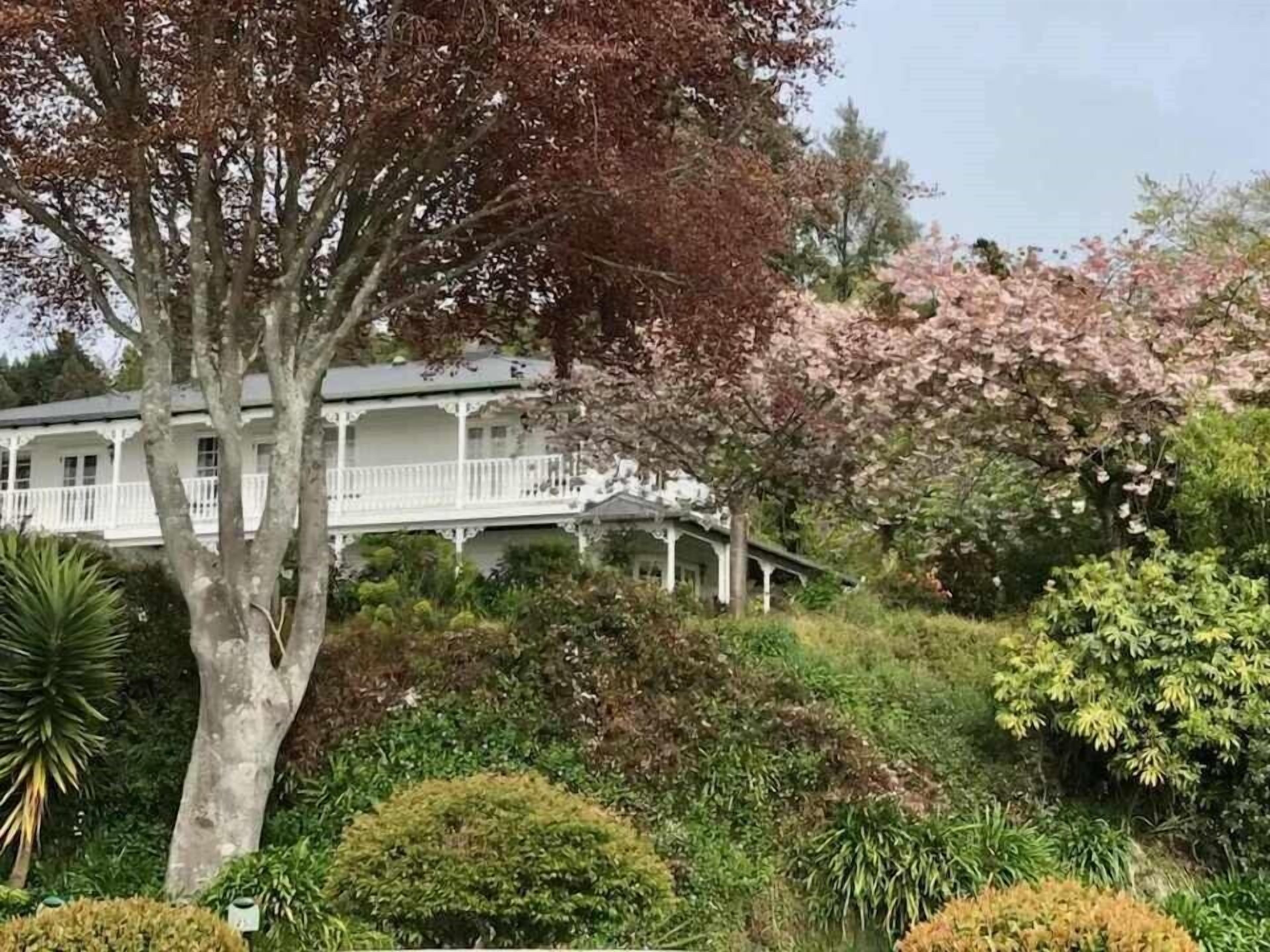 Ein weißes Haus im Kolonialstil mit einem Balkon, umgeben von üppiger Vegetation und blühenden Bäumen.