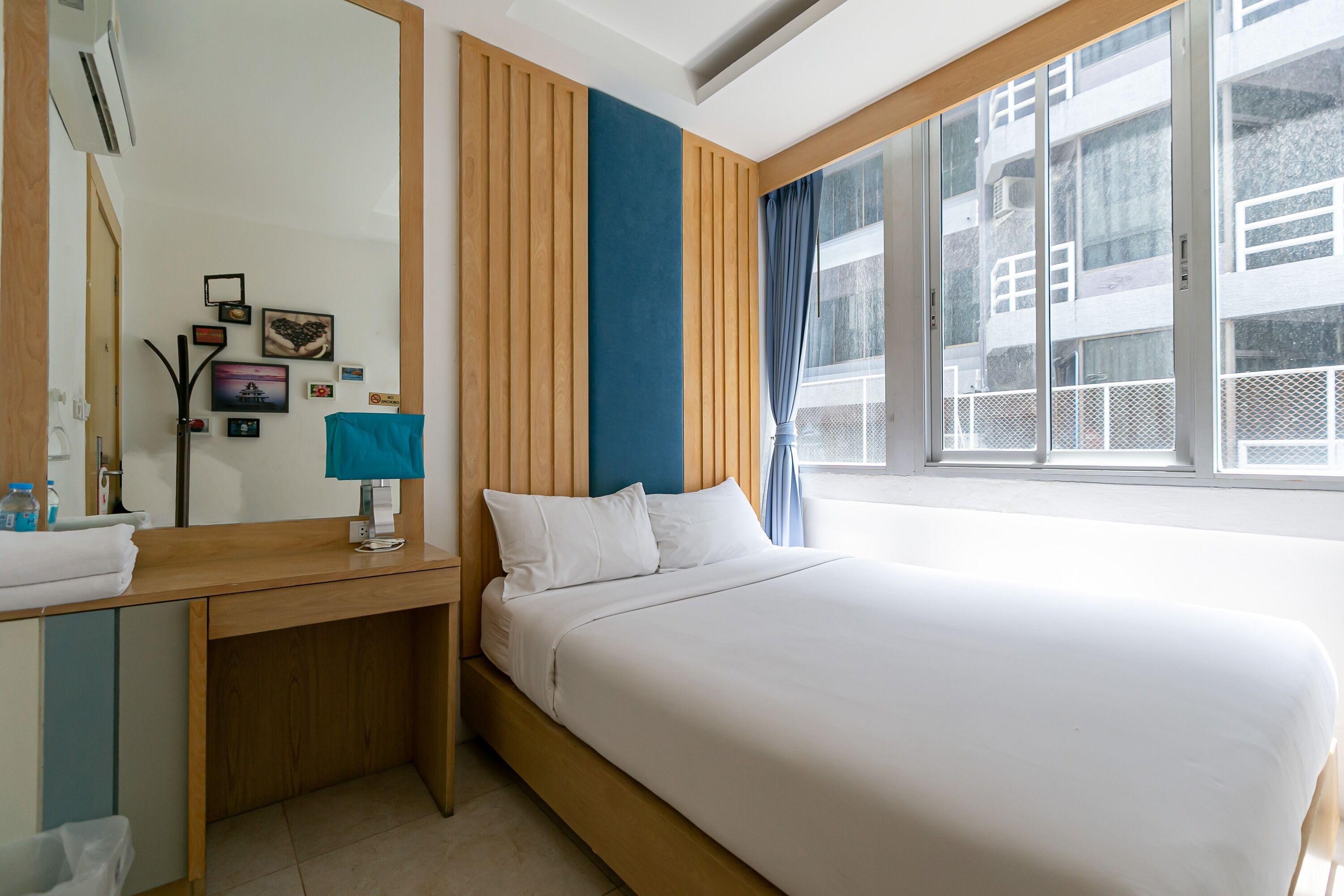 A hotel room with a large bed, a wooden desk, a mirror, and a window with a view of buildings.