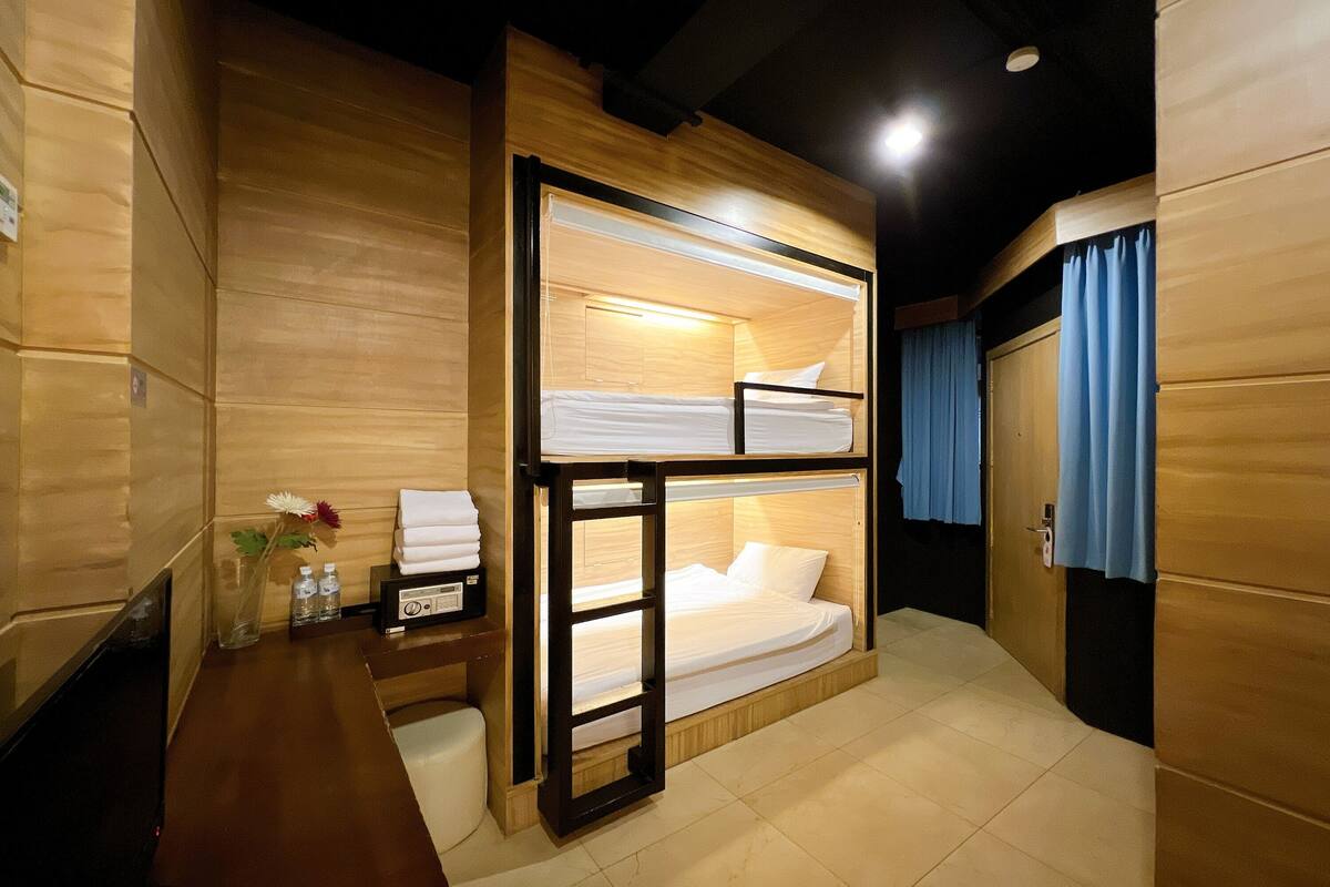 Een slaapkamer met stapelbedden, houten wanden, een nachtkastje met een vaas en flessen, en een blauw gordijn bij de deur.