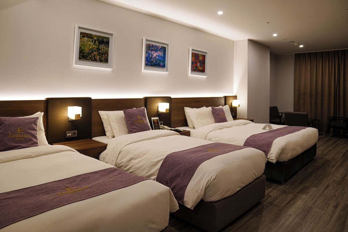 A hotel room with three beds, each with a headboard, bedside lamps, and a purple bedspread.