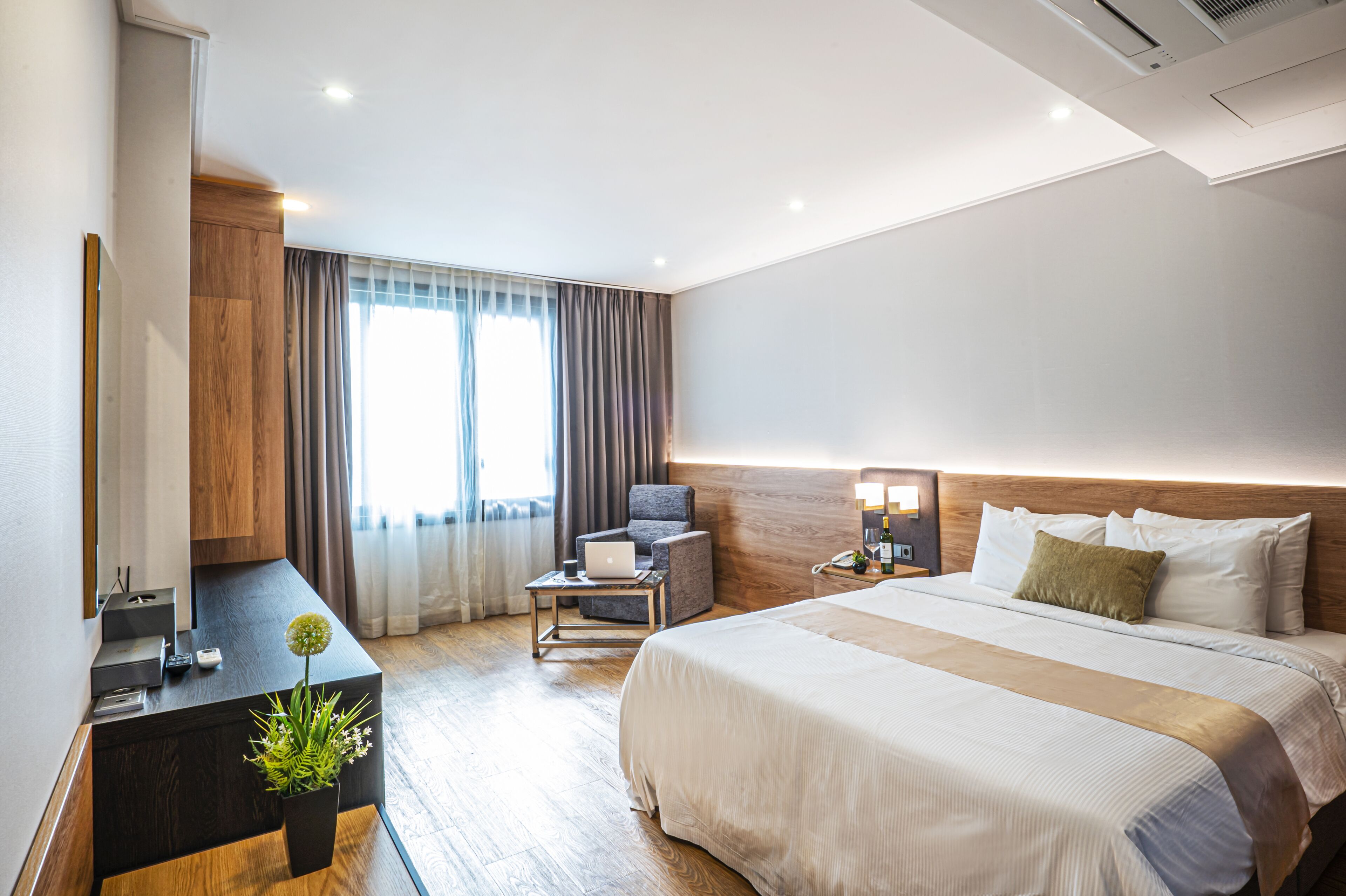 Habitación de hotel con cama, escritorio con computadora portátil, silla, mesita de noche con lámpara y planta en maceta.