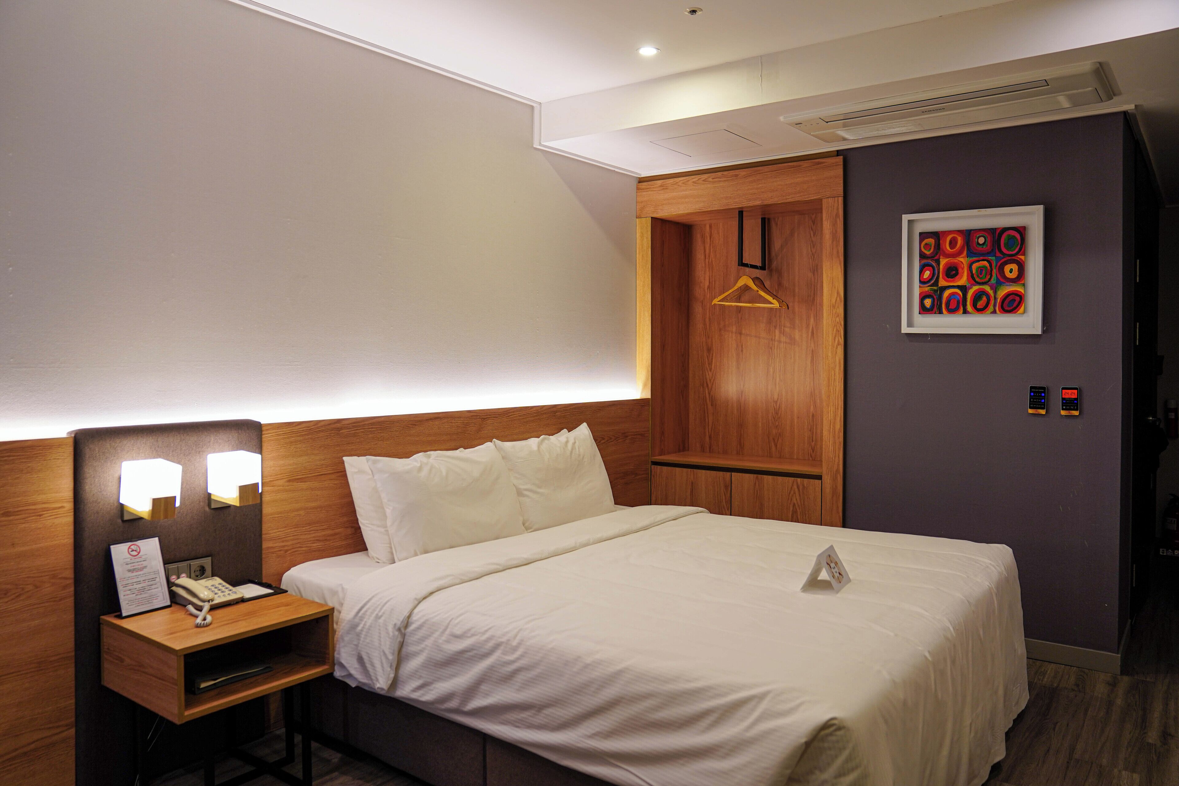 A hotel room with a bed, bedside table, lamp, and a framed picture on the wall.