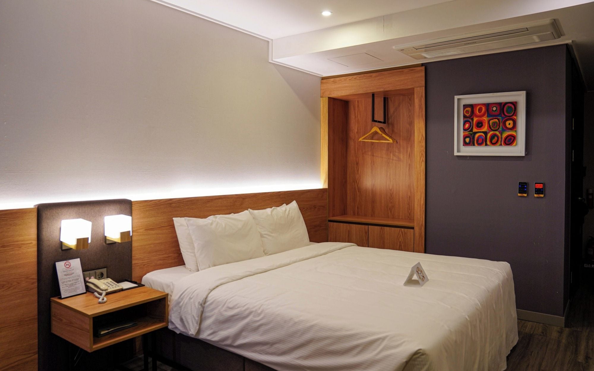 Camera d'albergo con un letto, un comodino con il telefono, un armadio in legno, una lampada a parete e un'opera d'arte incorniciata.