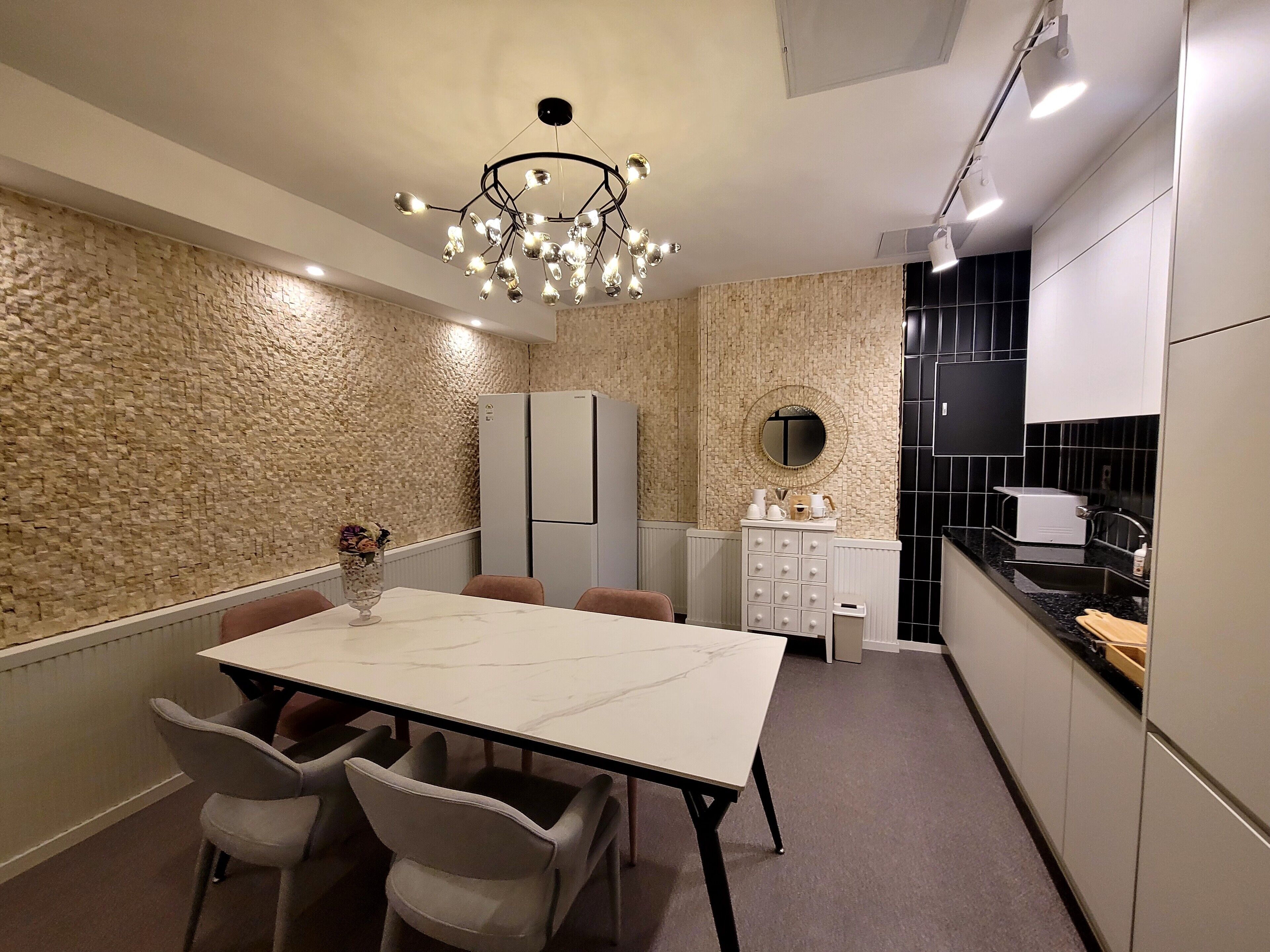 A compact kitchen with a dining area, featuring a marble table, grey chairs, a modern chandelier, and a built-in refrigerator.