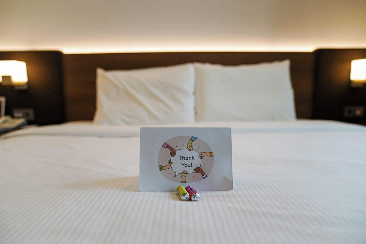A neatly made bed with white linens, a thank you card with a graphic of hands, and two small decorative items on the bed.