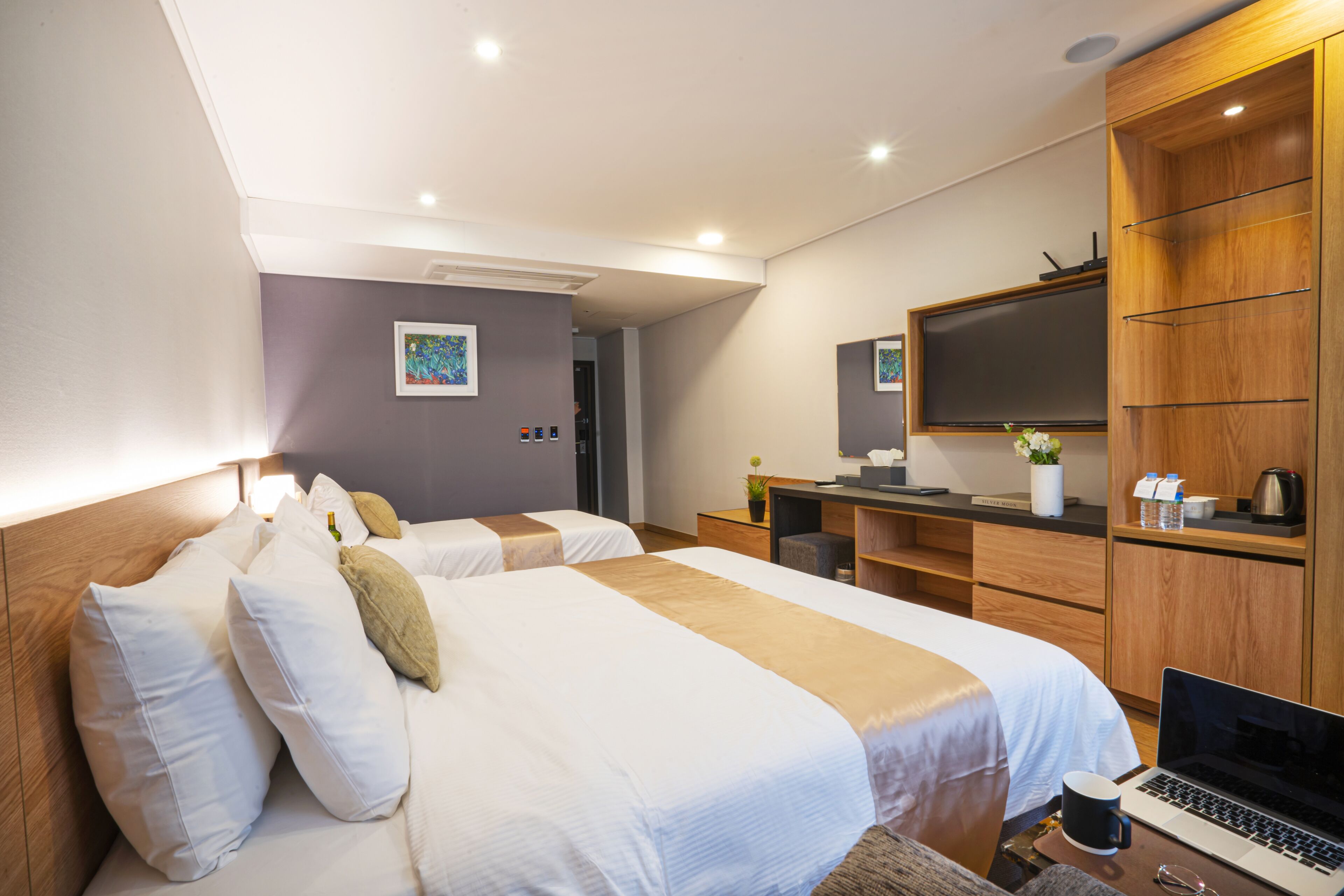 Una habitación de hotel moderna con dos camas, un escritorio con una computadora portátil, un televisor de pantalla plana y un armario de madera.