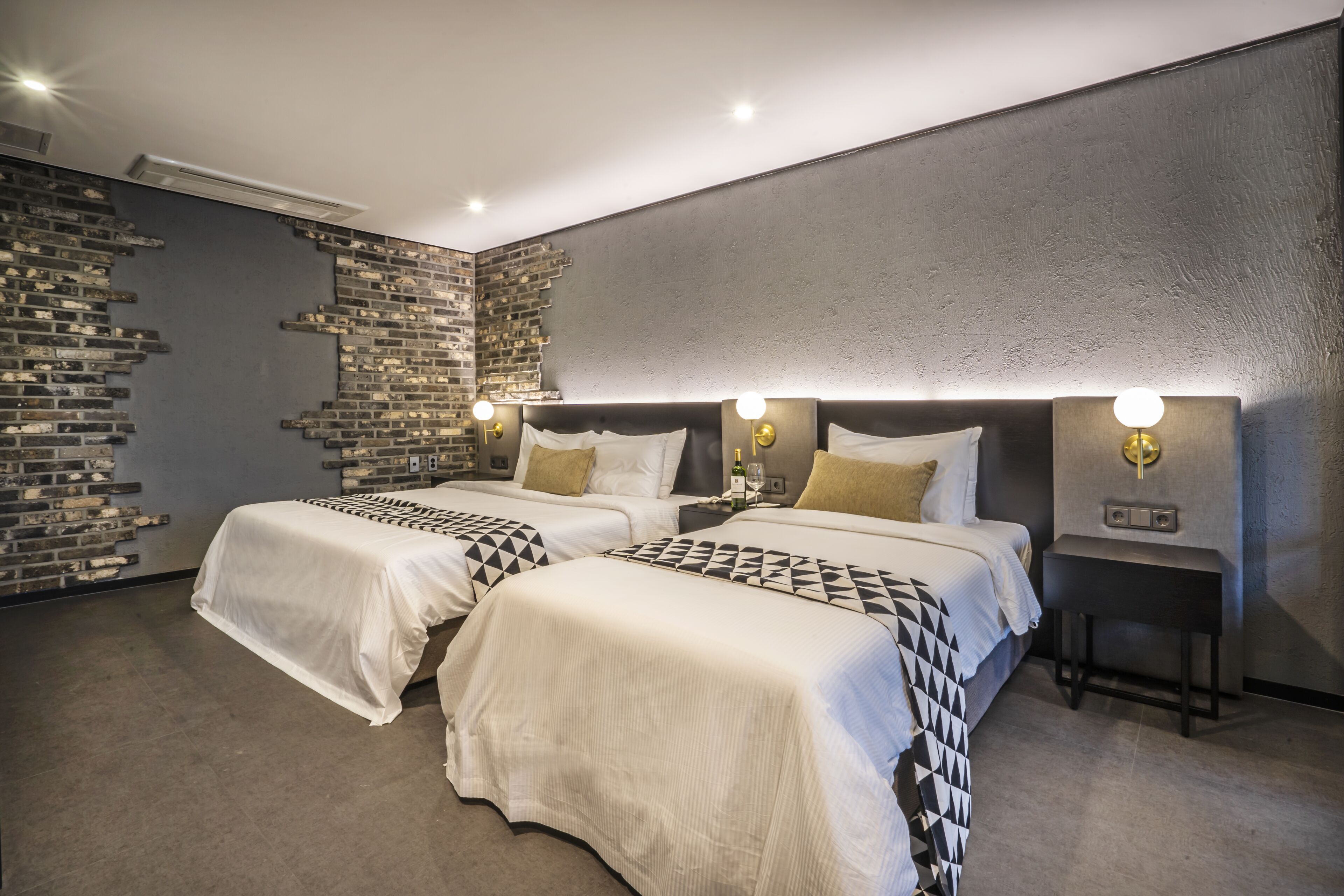 A modern hotel room with two beds, a brick accent wall, and a small bedside table with a lamp.