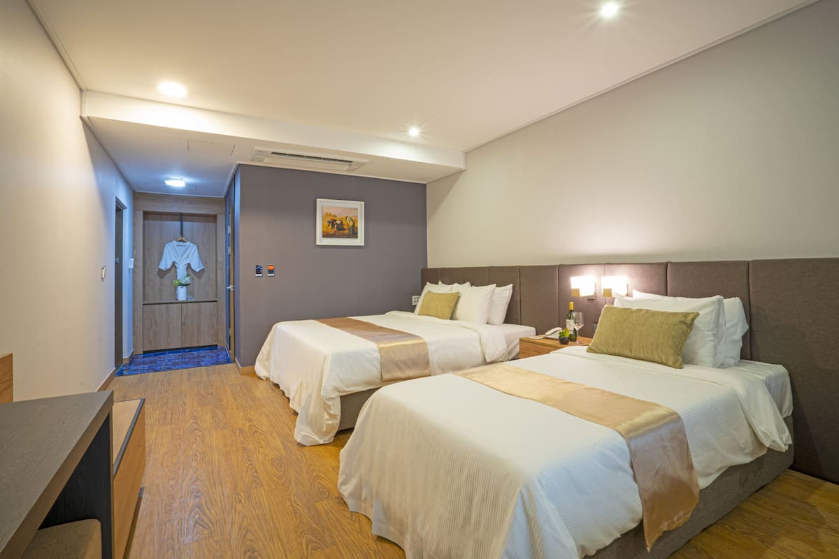 Quarto de hotel com duas camas, uma escrivaninha, uma cadeira, mesa de cabeceira e um quadro na parede.