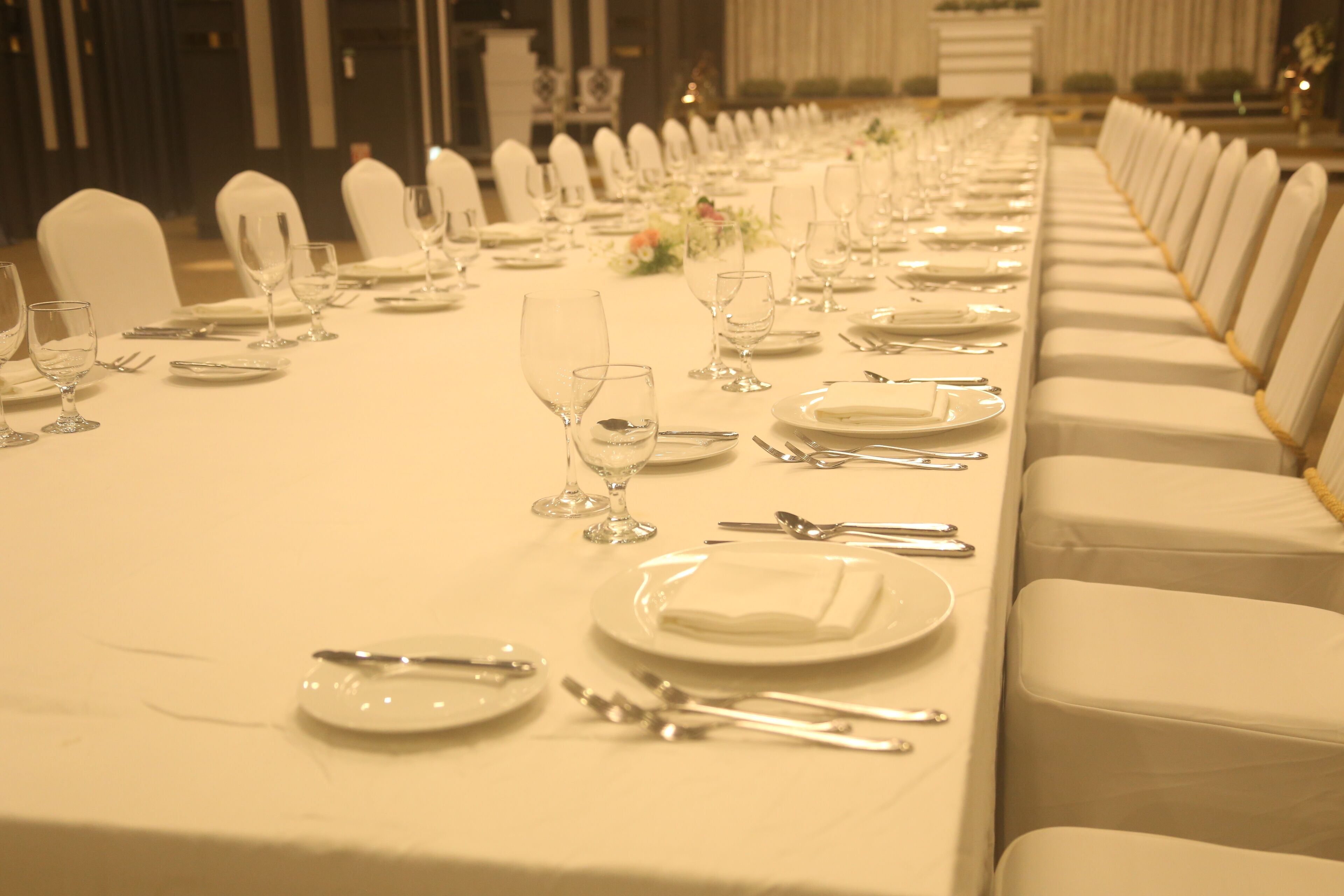 Una larga mesa de banquete dispuesta con sillas blancas, platos, cubiertos y copas de vino.