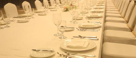 Una larga mesa de banquete dispuesta con sillas blancas, platos, cubiertos y copas de vino.