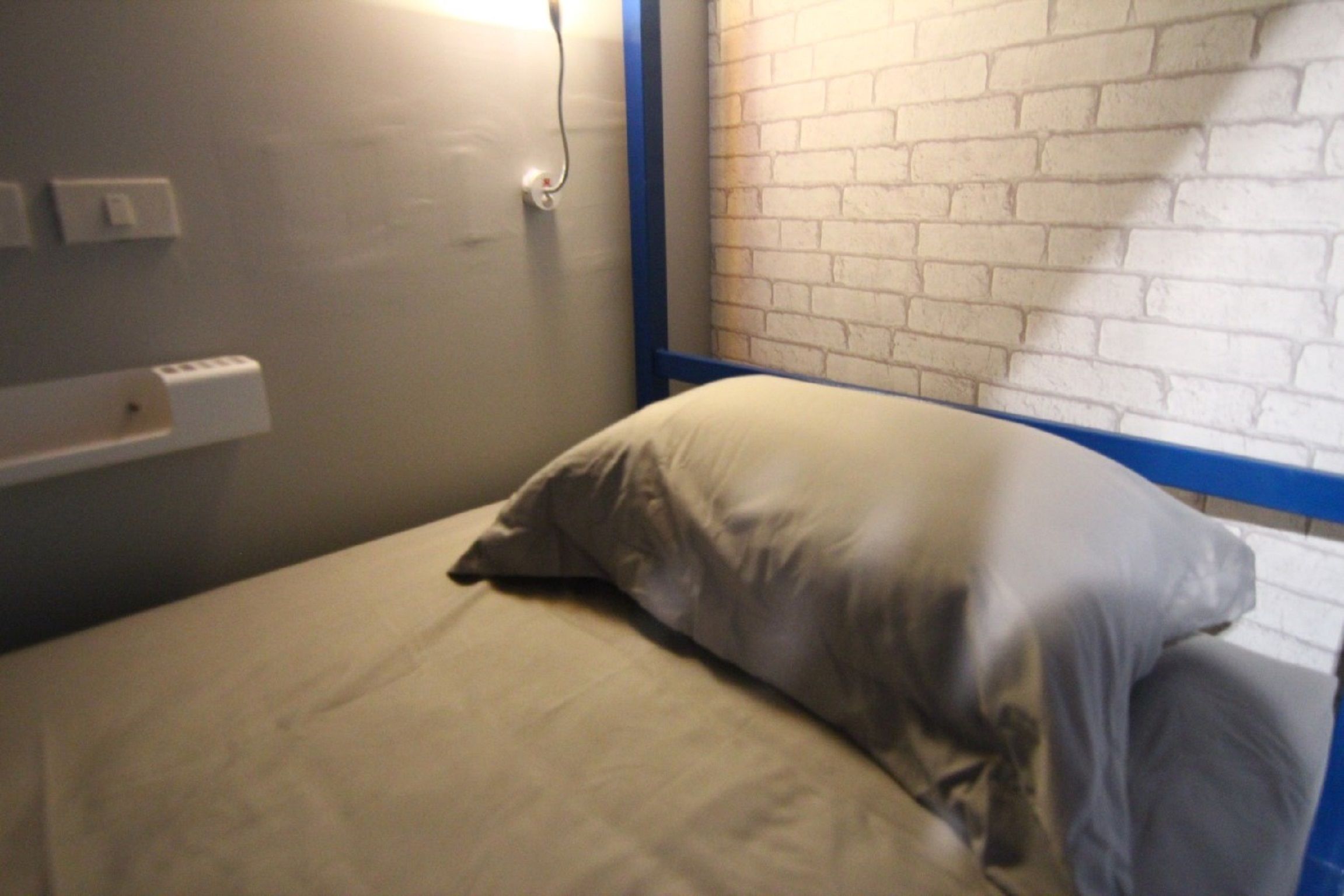 A single bed with a white mattress and a gray pillow in a room with a brick-patterned wall and a blue trim.