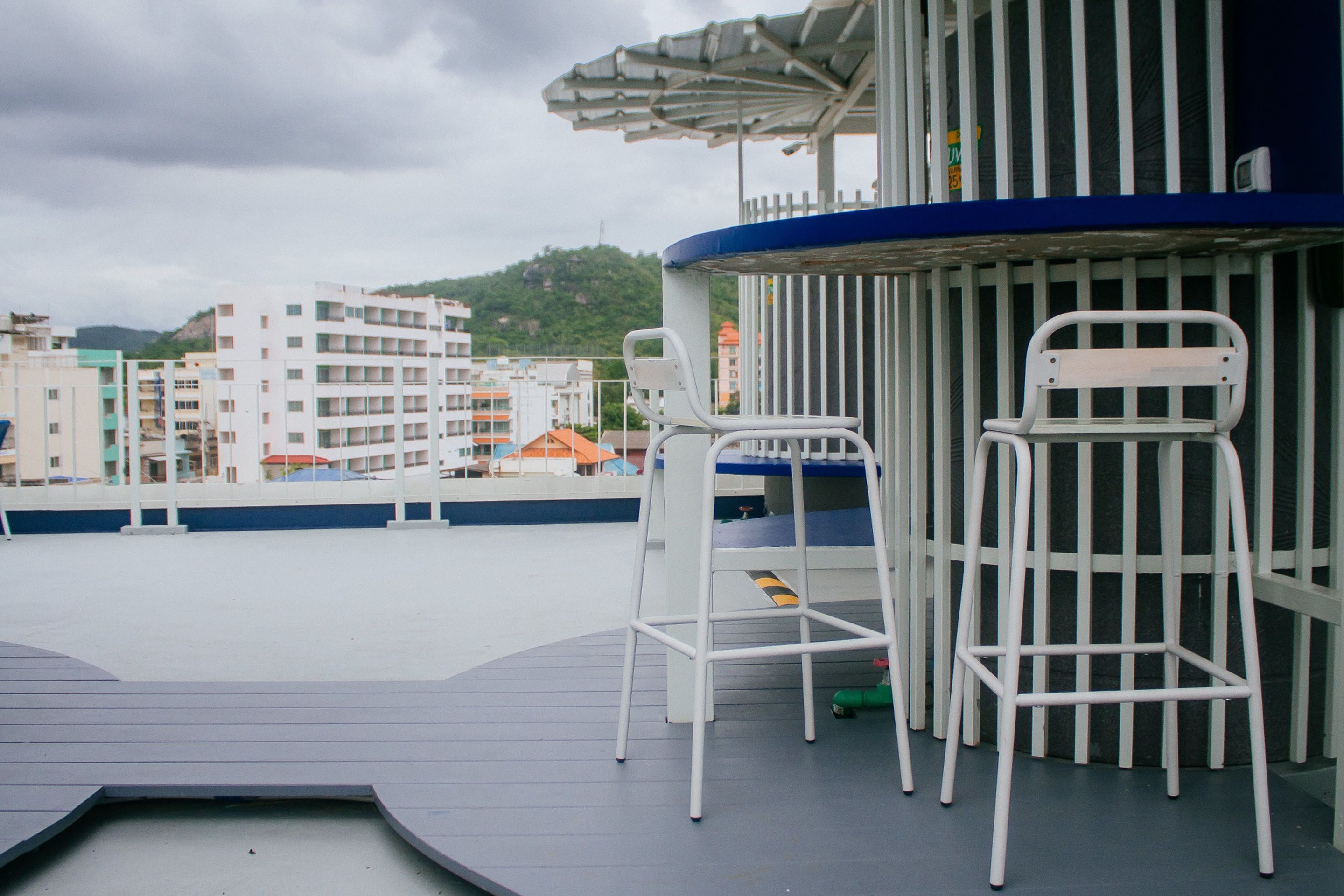 A rooftop terrace with white metal chairs and a small table, overlooking a cityscape with buildings and a hill.