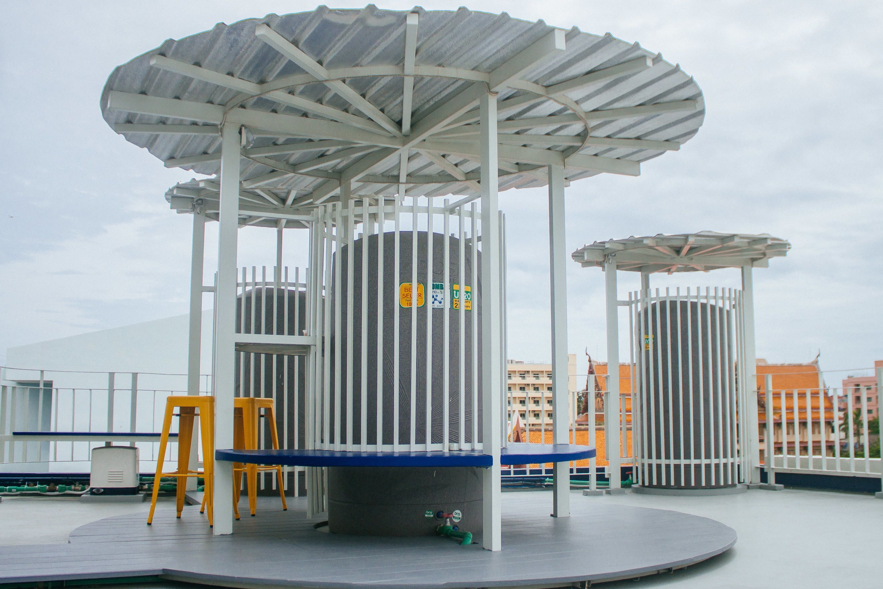 A covered outdoor seating area with a circular table and chairs, surrounded by white railings and a metal roof structure.