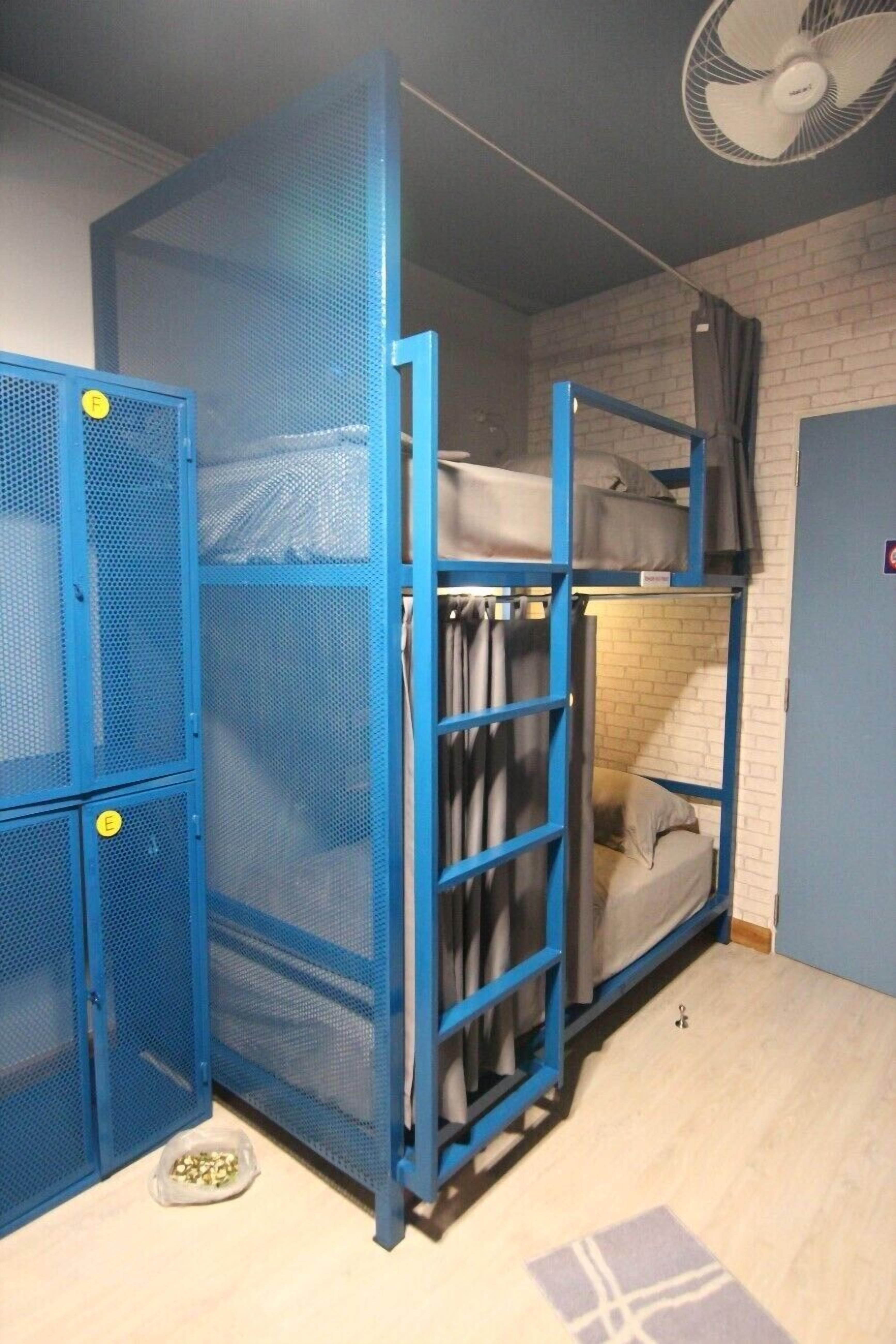 Uma cama beliche com estrutura metálica azul, um espaço pequeno fechado com cortina e um ventilador no teto.