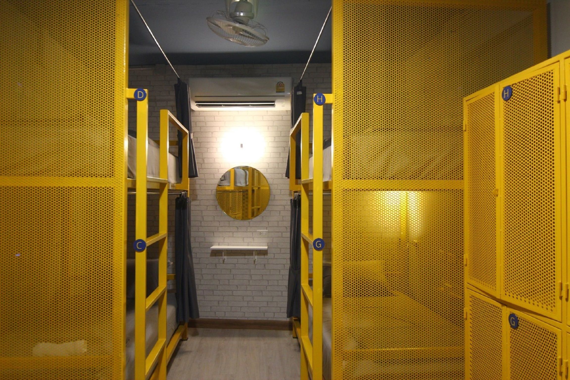 A narrow hallway with yellow lockers on both sides, a round mirror on the wall, and a white brick wall.
