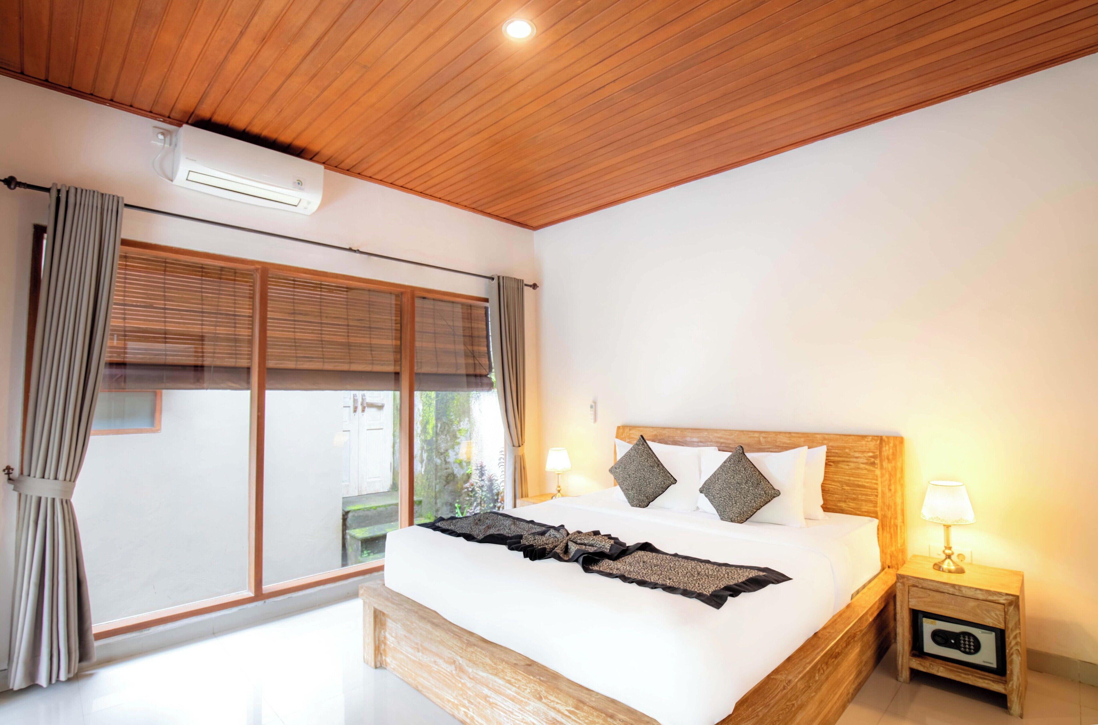 A bedroom with a wooden ceiling, a large window with curtains, a bed with a headboard, and a bedside table with a lamp.