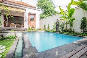 A rectangular swimming pool with a stone border, surrounded by a garden with various plants and a statue. - Sari Bamboo Villas (Ubud)