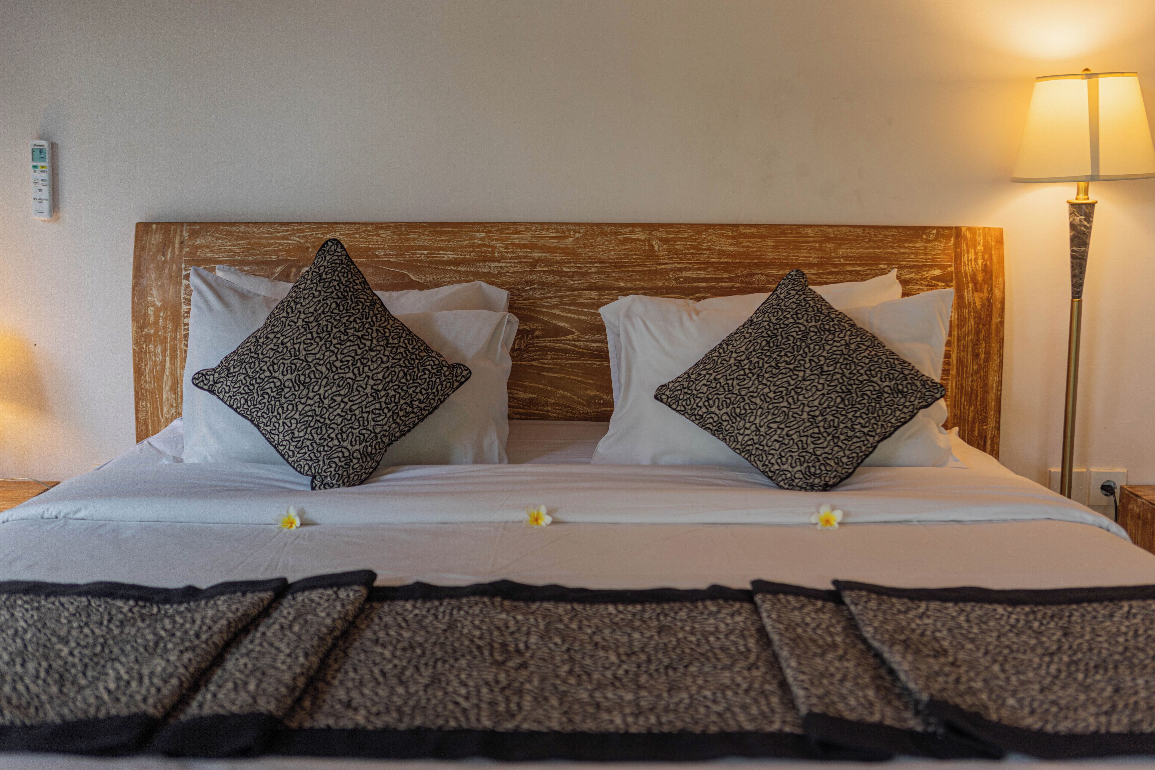 Una cama bien tendida con cabecera de madera, dos almohadas estampadas y una lámpara de noche.