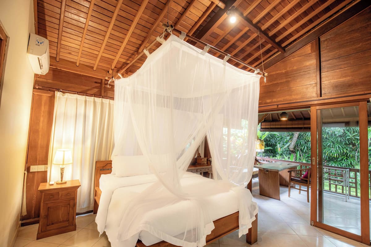 A bedroom with a wooden ceiling, a bed with a white canopy, a bedside table with a lamp, and a view of the outdoors through a sliding glass door.
