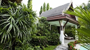 Un espacio exterior techado con piscina, rodeado de abundante vegetación y un edificio tradicional con tejado inclinado.