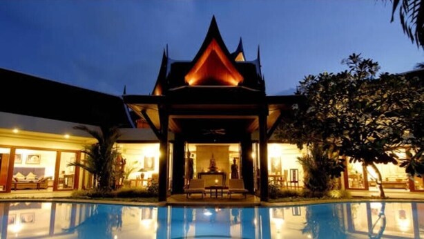 Un hotel con piscina al frente, una estructura arquitectónica tradicional con techo puntiagudo al fondo y una entrada bien iluminada.