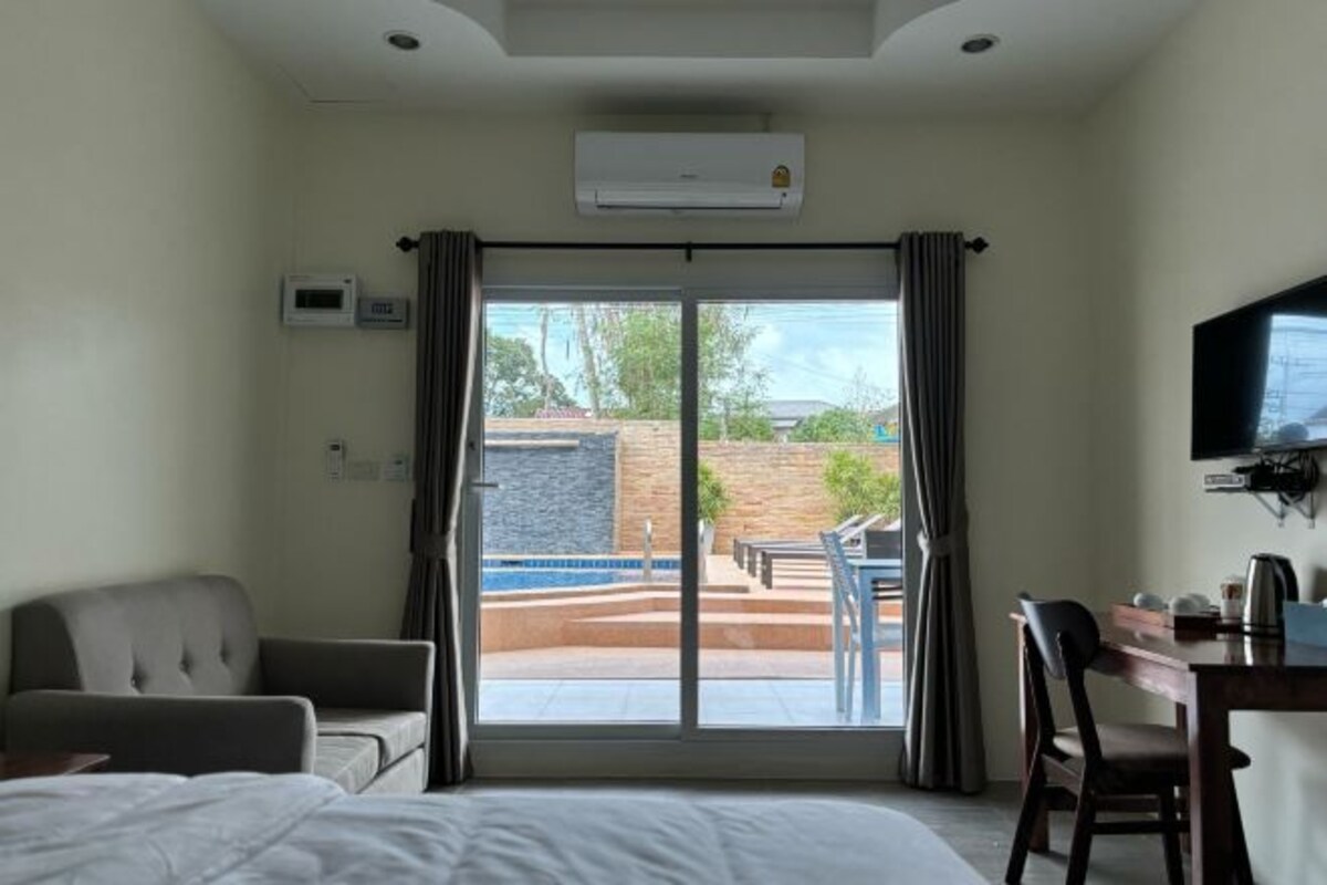 Habitación de hotel con cama, escritorio, silla, equipo de aire acondicionado y puerta corrediza de vidrio que da a un área exterior con piscina y mobiliario de patio.