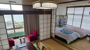 A hotel room with a large window, a bed with a white and pink duvet, a dining table with a remote control, and a red chair.