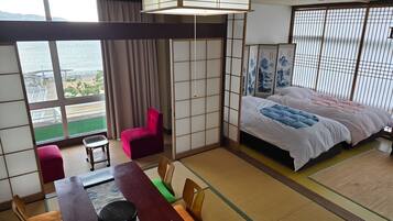 Une chambre d’hôtel avec une grande fenêtre, un lit recouvert d’une couette blanche et rose, une table à manger avec une télécommande, et une chaise rouge.