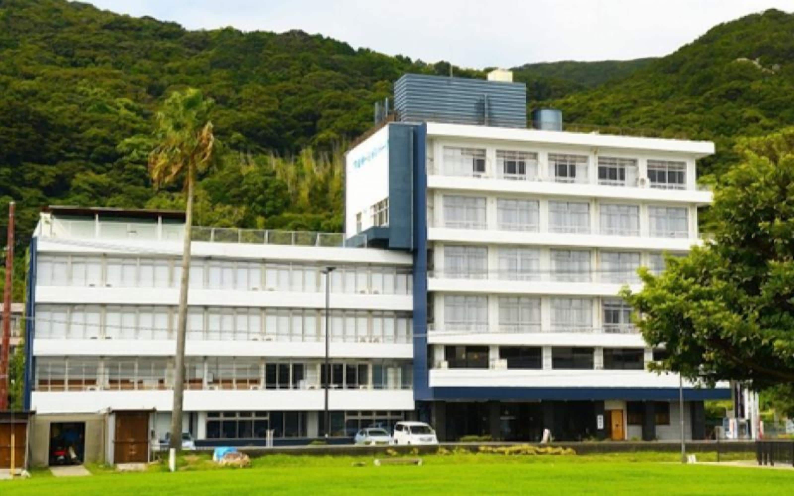 A multi-story building with a flat roof, surrounded by greenery and hills.