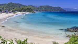 A beach with clear turquoise water, sandy shore, and green hills in the background.