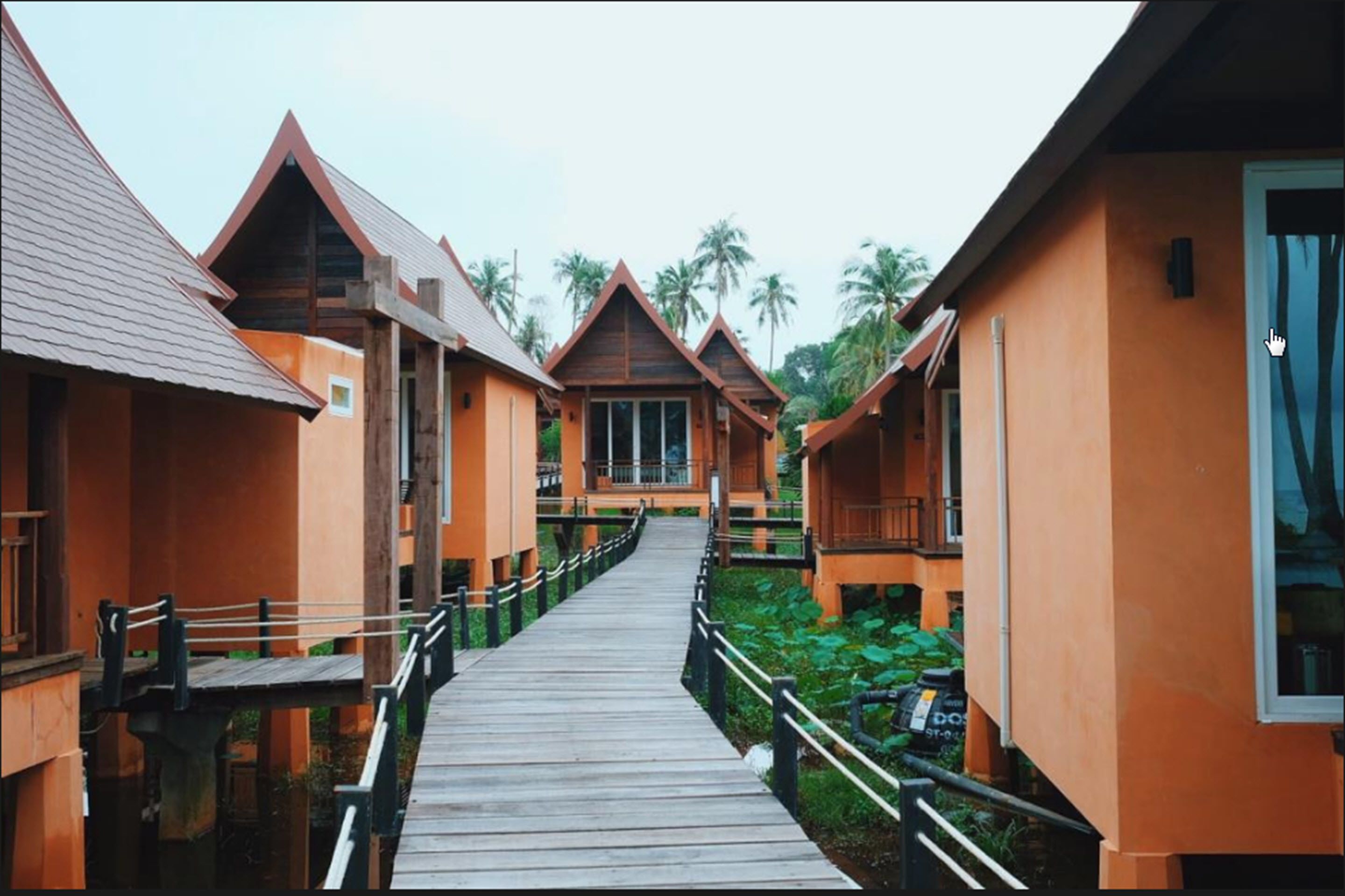 A wooden pathway leading to a row of orange-roofed cottages with balconies, surrounded by greenery.