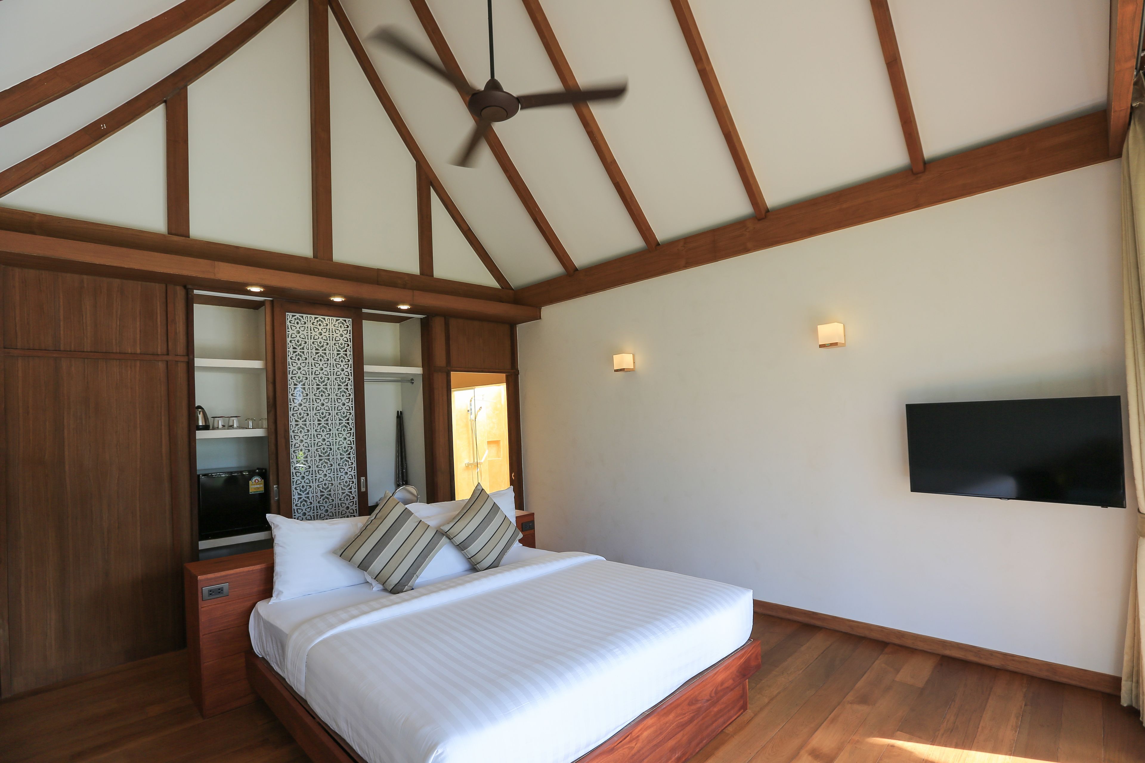 Un dormitorio con techo de madera, cama con ropa de cama blanca, televisor de pantalla plana y ventilador de techo.