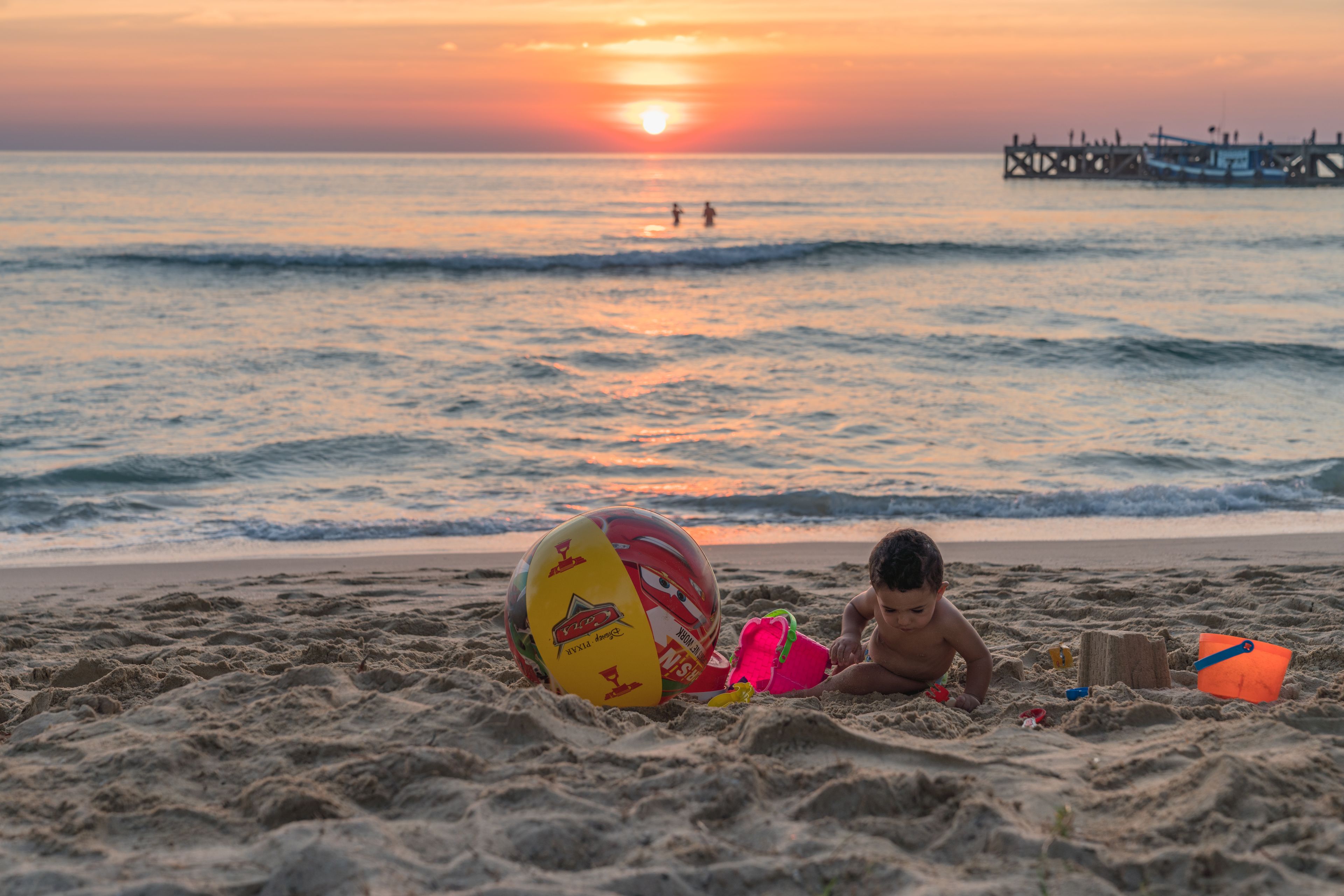 Une scène de plage avec un enfant qui joue dans le sable, un ballon coloré et une jetée en bois en arrière-plan.
